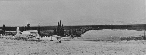The Lerna mound from the west, with a small church and white buildings among cypresses.