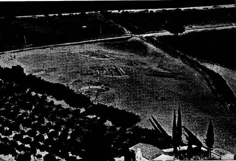 Telephoto view from Mt Pontinos onto the Lerna excavation, with in the foreground a small church with bell tower and cypresses and roofs of Myloi, an olive grove, the road/railway and the WWII tank trap.
