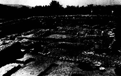 Part of the stone circle around the remains of the House of the Tiles at Lerna, with trees and hills behind.