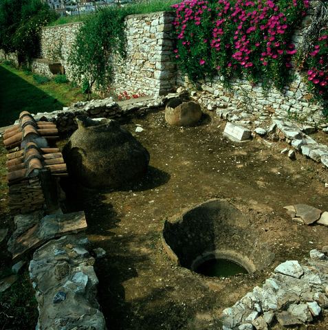 Lerna excavation: pithoi and a bothros, a dry-stone wall with flowering bougainvillea/oleander, and houses behind.