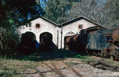 'Mili Nafpliou Depot. Greece. 1995.': the stone front of the double engine shed with two gables, three arched doors and two round brick-edged windows, with steam locomotives (Di 7112, 7118, Es 7728) in and beside the doors.