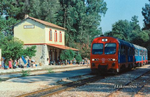 Colour photo of Myloi station in use: the SPAP station building with the ΜΥΛΟΙ sign and canopy, many passengers with luggage on the platform, and OSE railcar 6529 arriving.