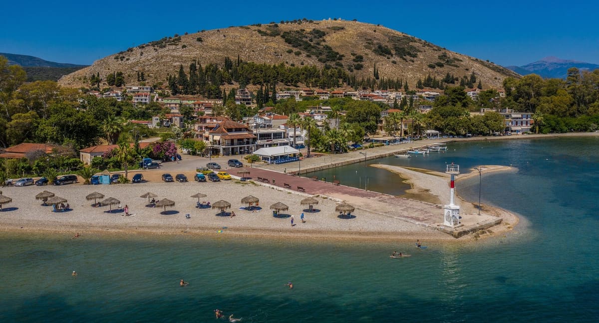 Aerial view of Myloi: pebble beach, pier with lighthouse and the waterfront, with Pontinos hill and its castle ruins behind the village