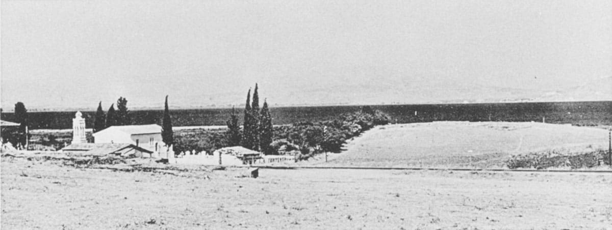 Black-and-white panorama of the Lerna mound from the west, with a white church, cypresses and the plain behind