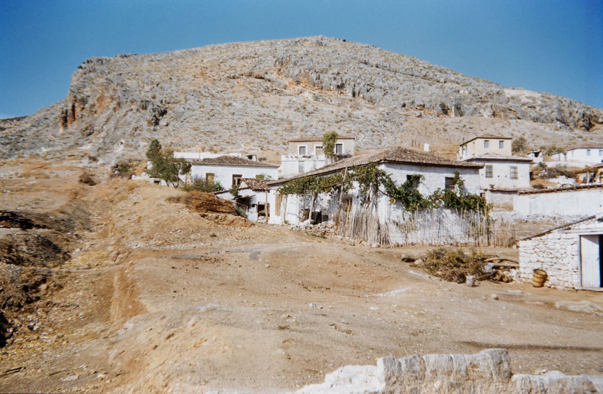 Whitewashed village houses with a vine-covered pergola and a reed fence at the foot of a rocky hillside in Myloi, 1950s