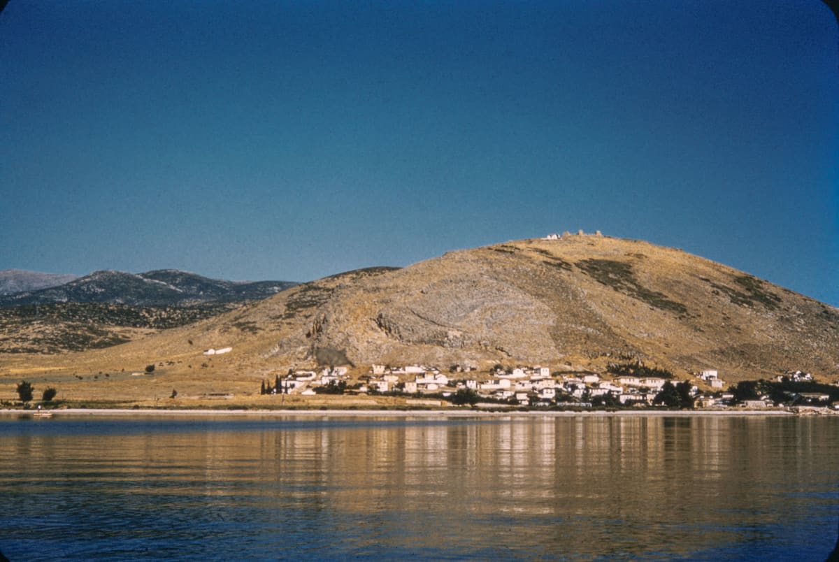 Myloi in the 1950s seen from the water: white houses along the shore at the foot of the bare hill of Pontinos, with the castle ruins on top, reflected in the calm gulf