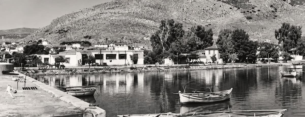 Black-and-white view of the harbour quay with rowing boats, white buildings and Pontinos hill behind (digitally restored)