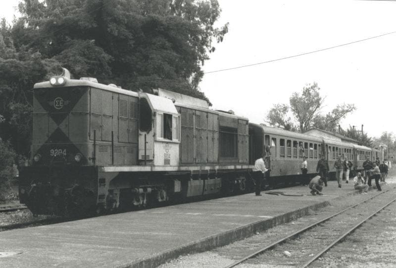 Black-and-white photo of a diesel locomotive and passenger train at Myloi station
