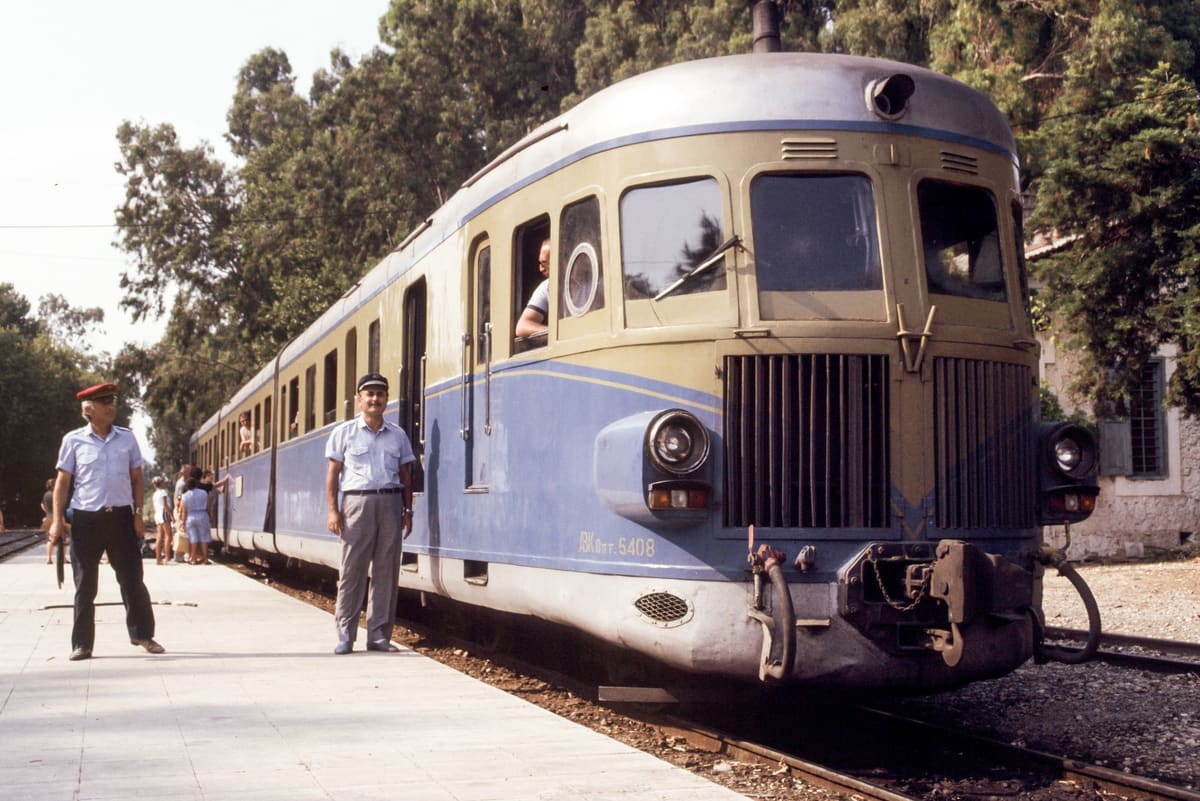 Front of a blue and beige railcar at Myloi with two railwaymen on the platform