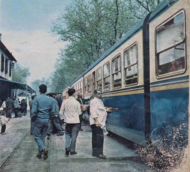Waiters in white jackets carry trays of souvlaki along a blue train at Myloi station