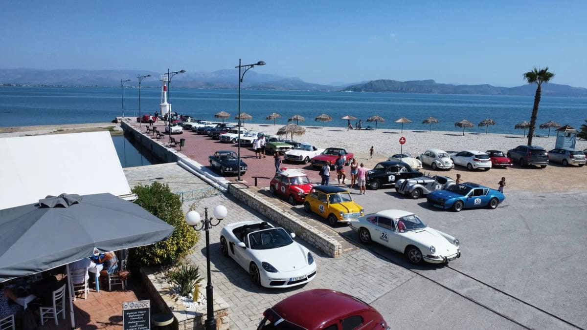 Classic rally cars with start numbers parked on the pier and waterfront of Myloi, seen from above, with the lighthouse and the gulf behind