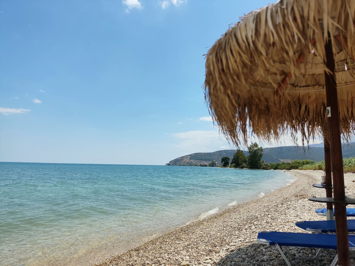 Clear turquoise water along the pebble beach, with a straw umbrella and blue sunbeds