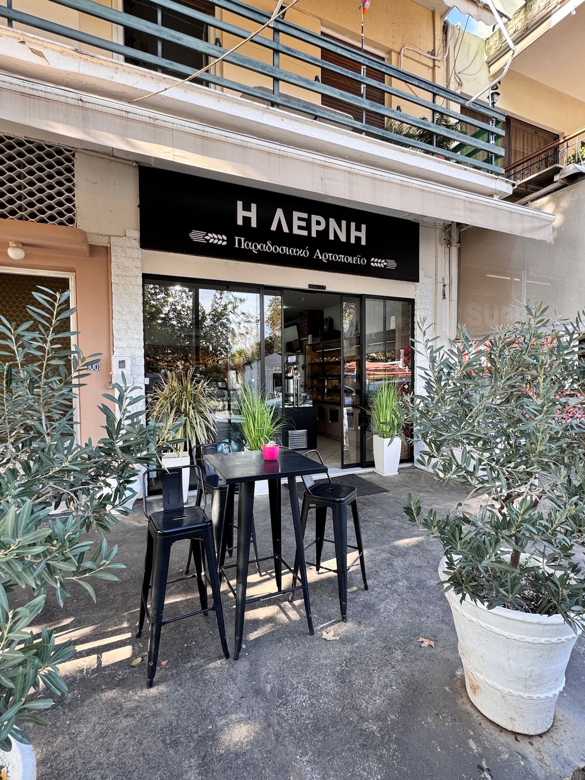 The front of the bakery I Lerni with its black sign, plants and high tables outside