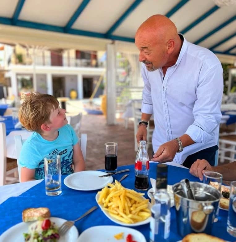 Kostas, owner of Faros, chatting with a young guest at a table under the taverna awning
