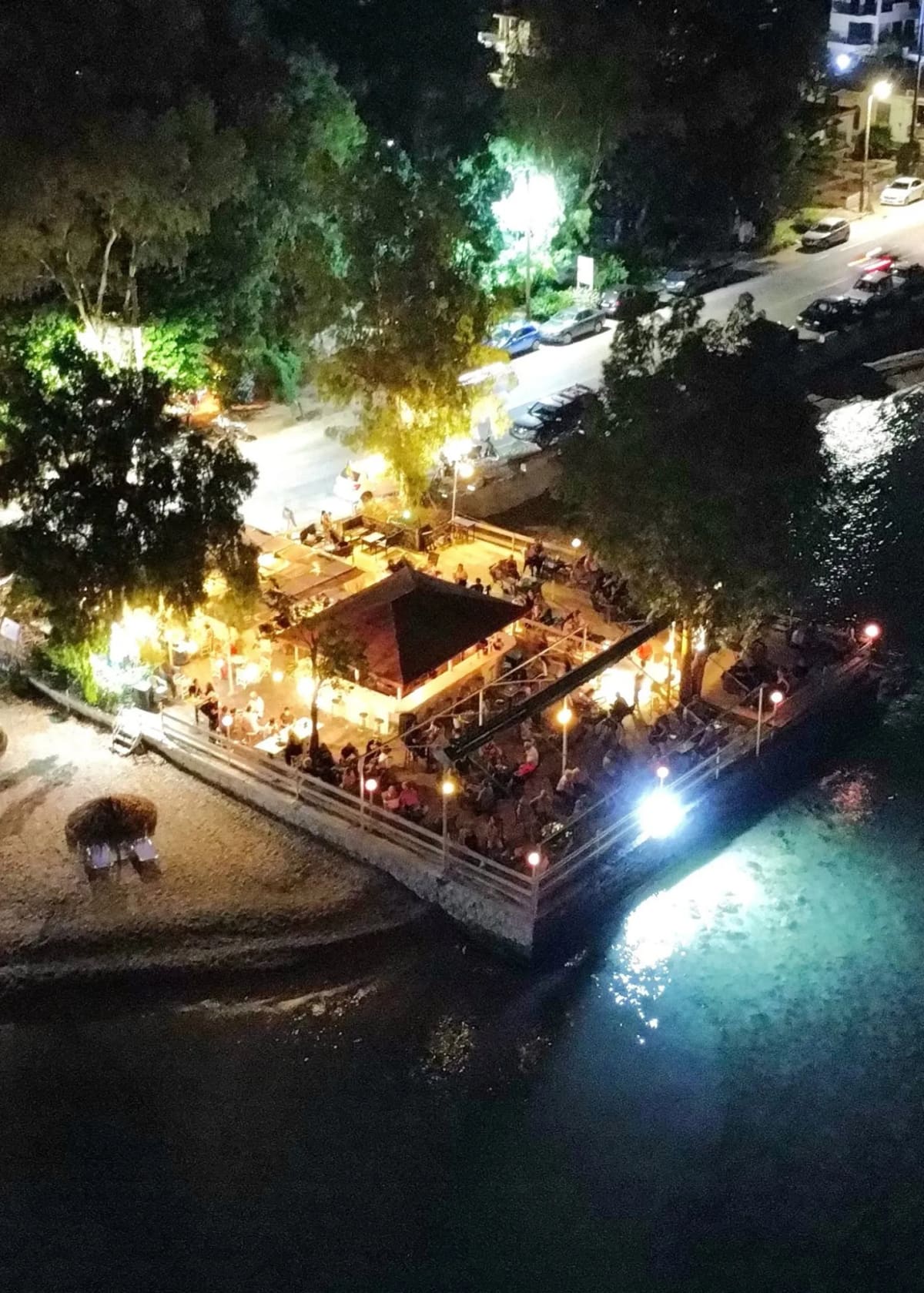 Grammes café-bar at night from above, a busy lit terrace at the water’s edge next to the beach