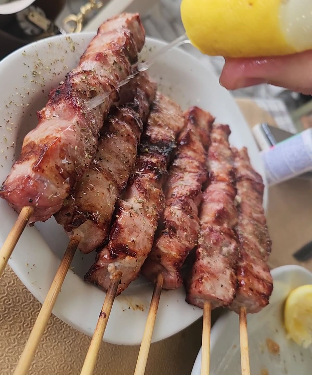 A plate of grilled pork souvlaki on wooden skewers, with a lemon being squeezed over
