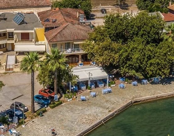 Lykos from the air: the taverna with its white awning and blue tables on the paved quay by the sea, palm trees and a large shady tree