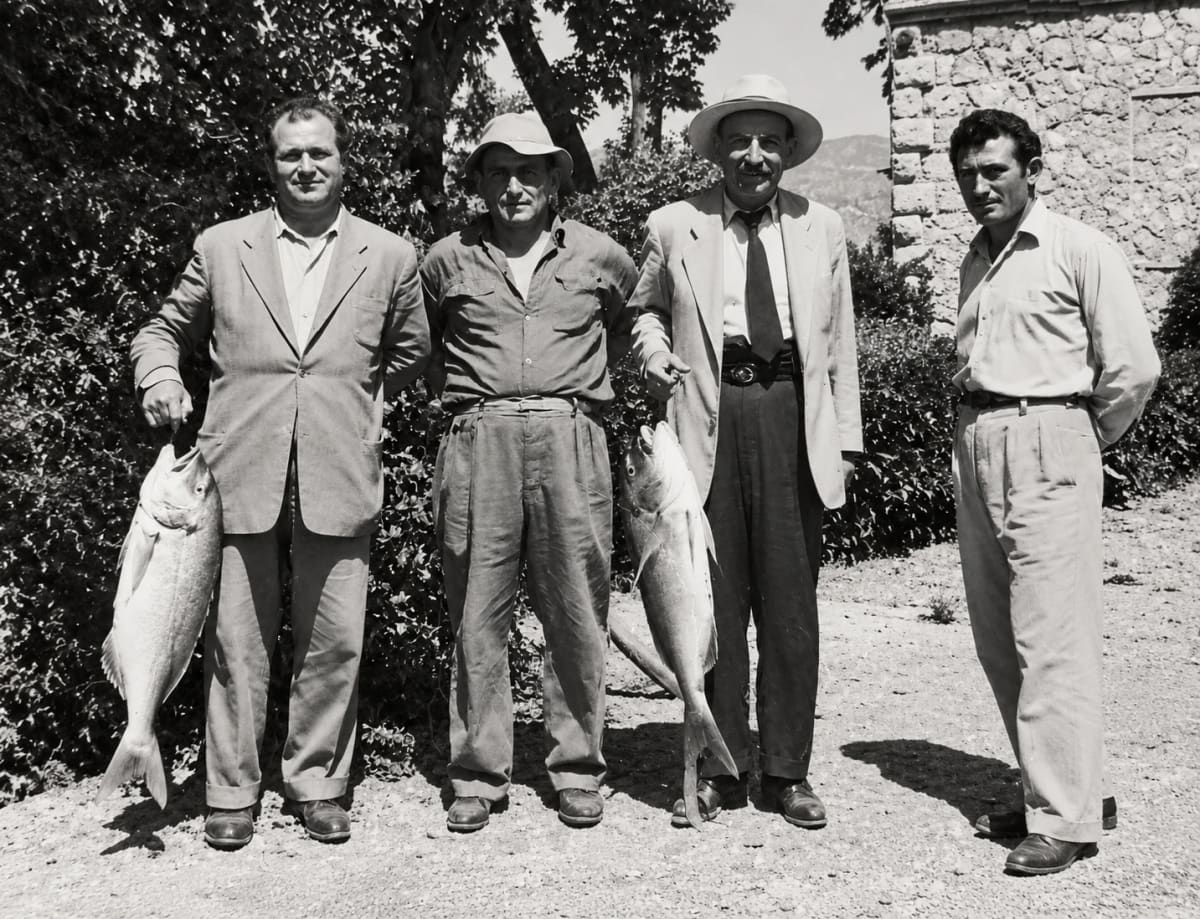 “The old fishermen of Myloi”: four men in front of a stone building, two of them holding large fish (digitally restored)