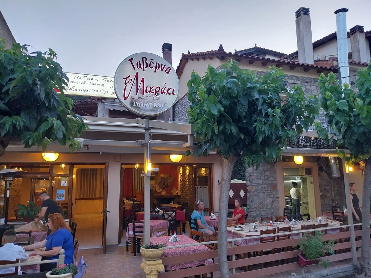 The front of the family taverna To Mikraki at dusk, with its round sign, warm lights and tables on the terrace
