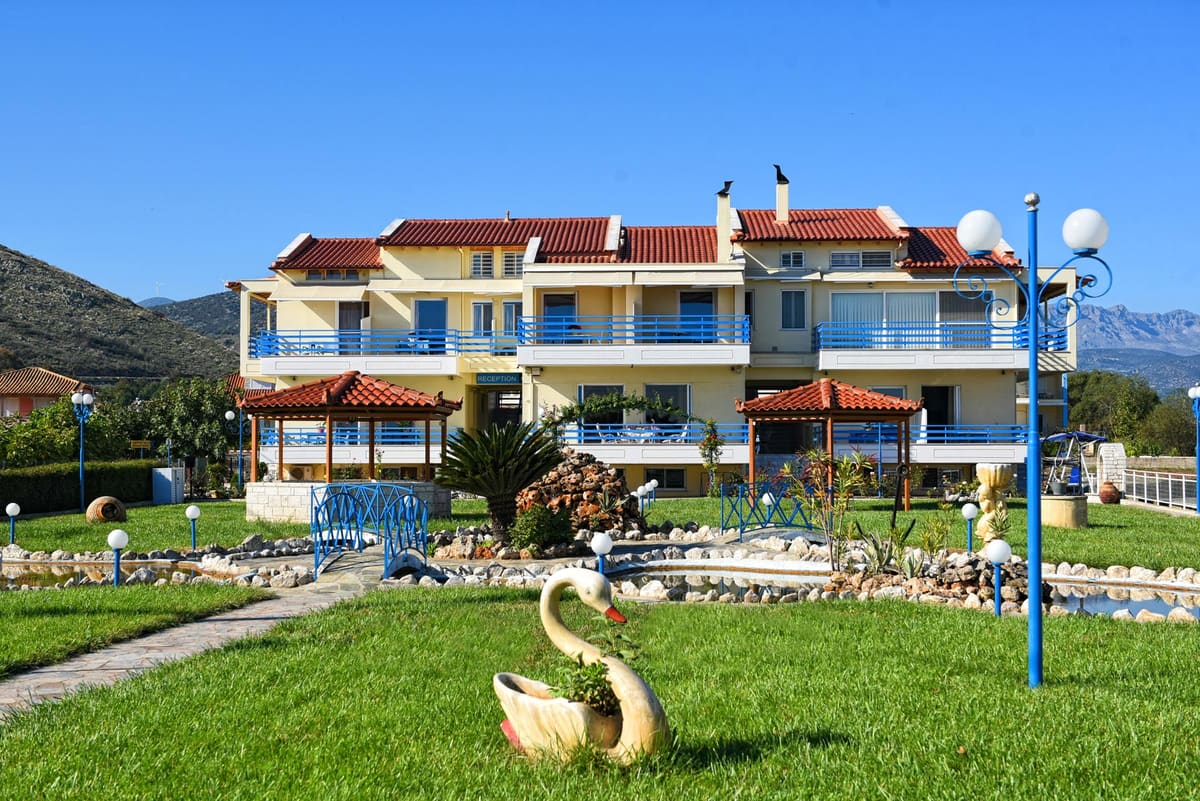 The two-storey Akrogiali building with blue balconies and red-tiled roofs, behind a lawn with a garden path and lamps