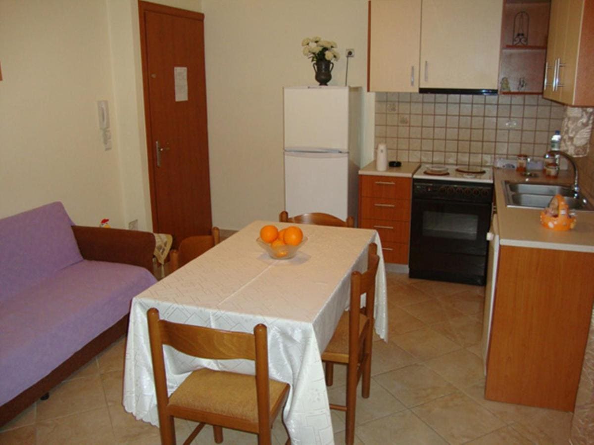 Apartment living area with a sofa, a dining table with a bowl of oranges, and a kitchen corner with oven and fridge