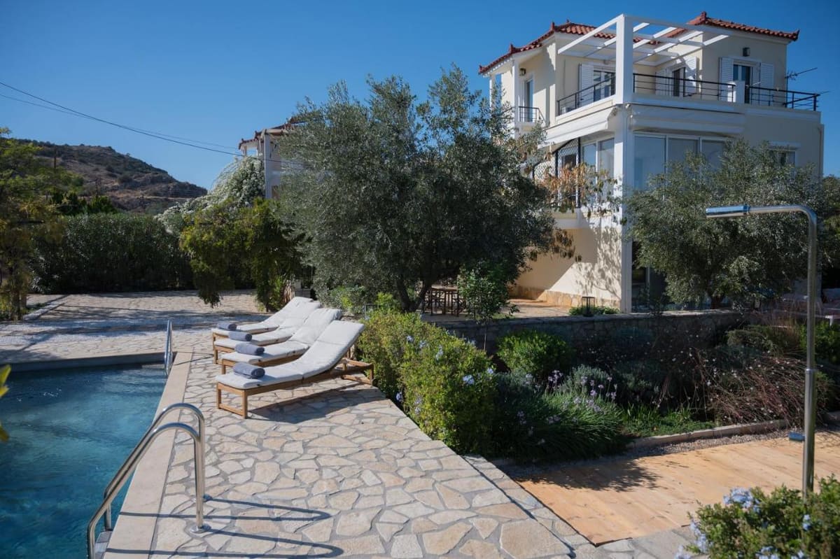 Swimming pool with white sun loungers on a stone terrace, gardens and olive trees around and the white villa behind