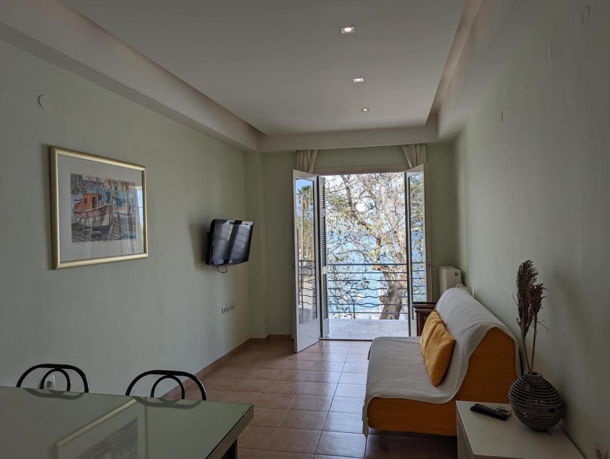 Apartment with a sofa, a dining table and the balcony door open to the trees and the sea
