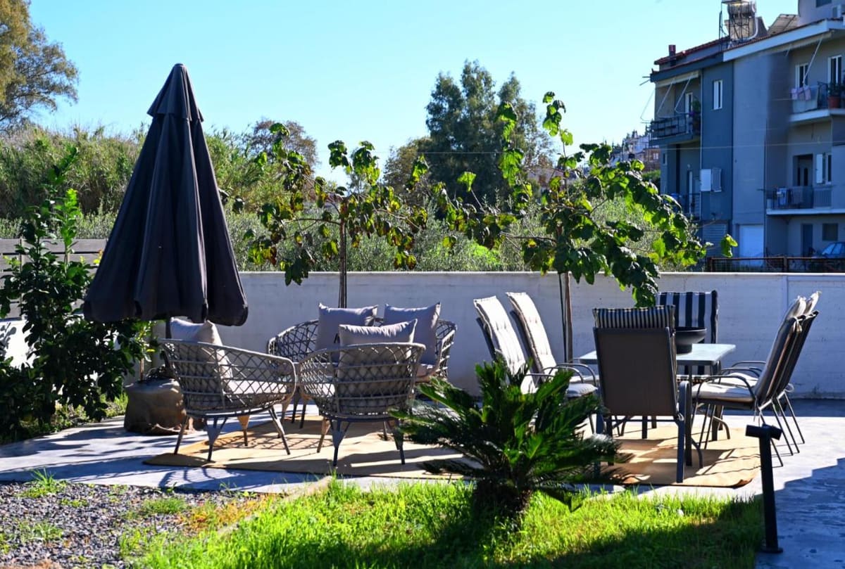 Sunny garden with an outdoor dining table, wicker chairs, a black parasol and plants along a white wall
