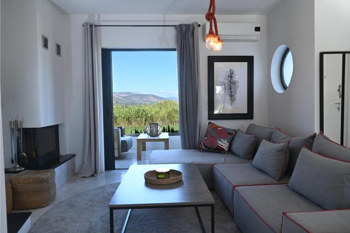 Modern living room with a large grey sofa, a corner fireplace and the glass door open to green fields and hills