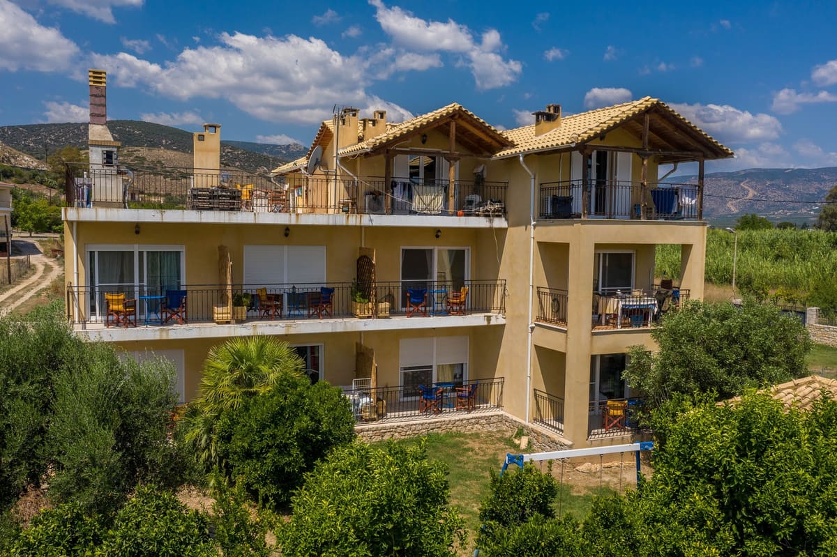 The ochre three-storey Myloi Inn with balconies and tiled roofs, behind a stone wall and olive trees