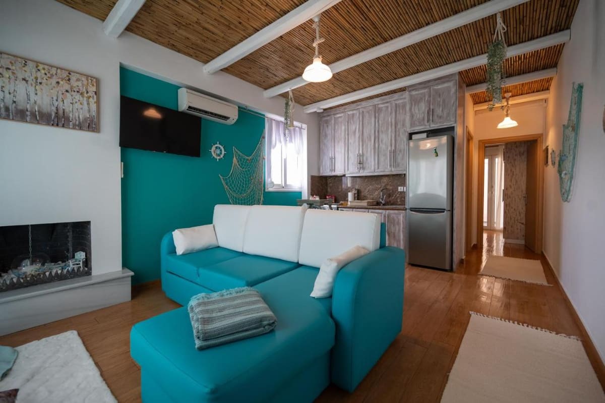 Living room in white and turquoise with a corner sofa, a fireplace, a reed ceiling and the kitchen at the back