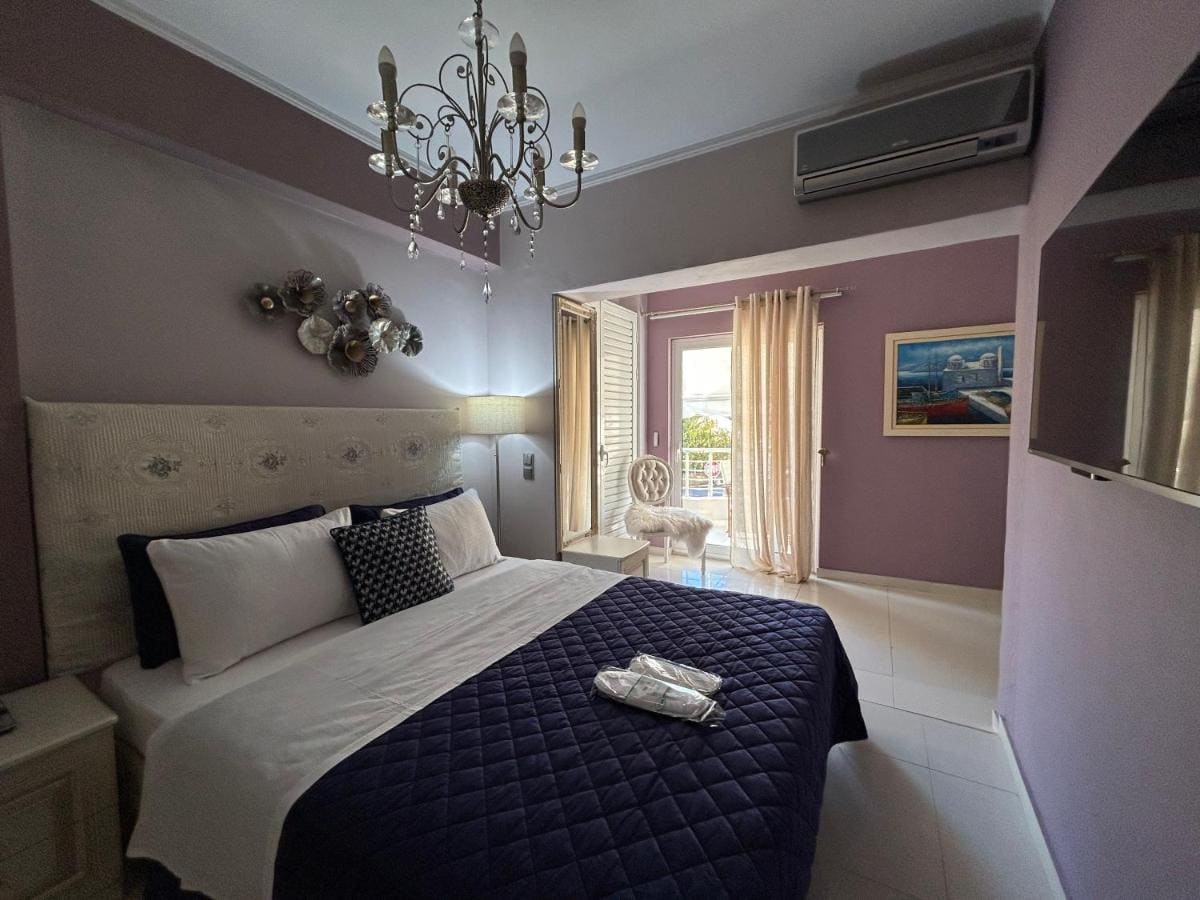 Bedroom with a crystal chandelier, a double bed with a navy quilt, lilac walls and a door to the balcony