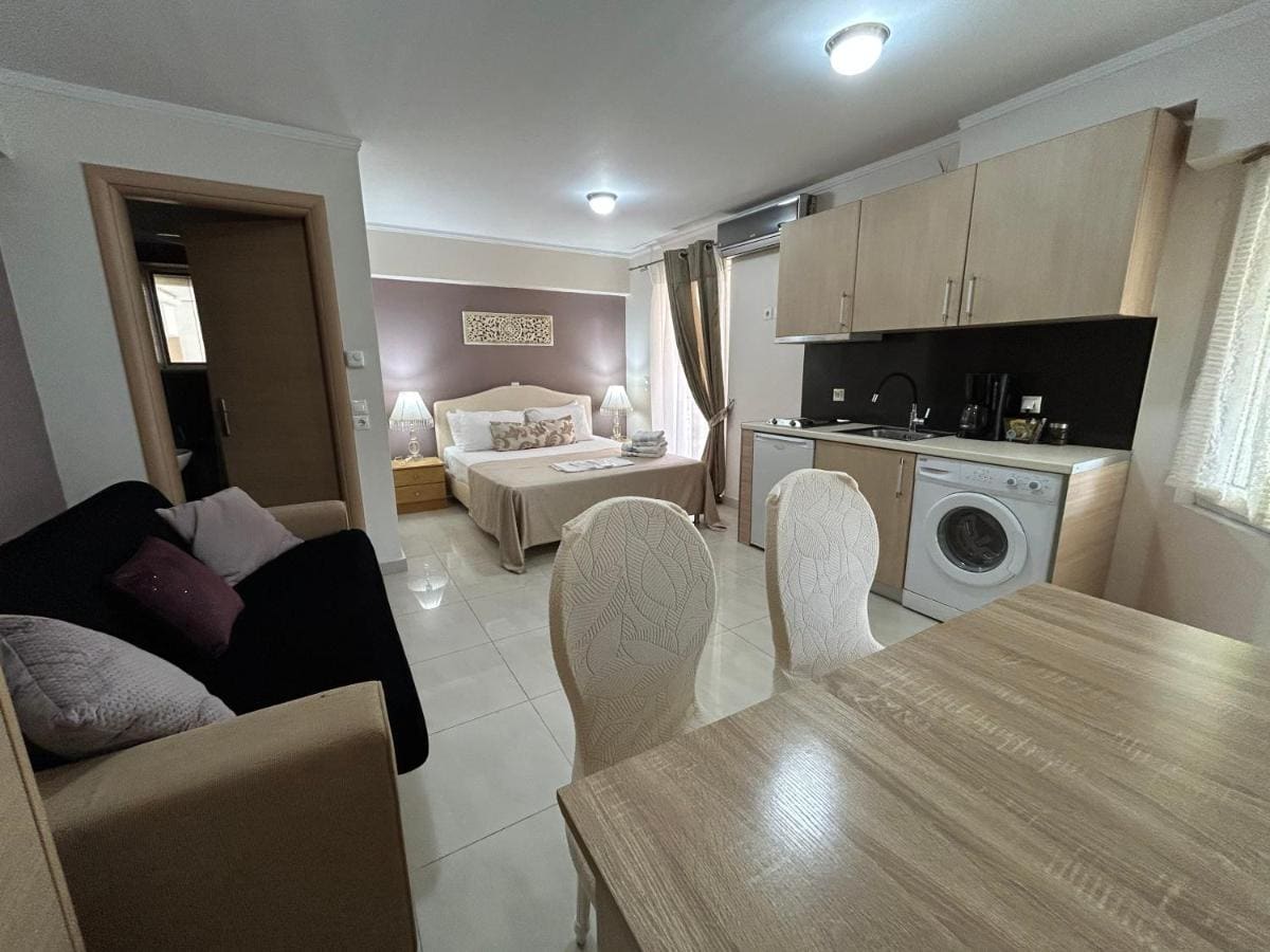 Open-plan apartment with a fitted kitchen and washing machine, a dining table with white chairs, a sofa and the bed beyond