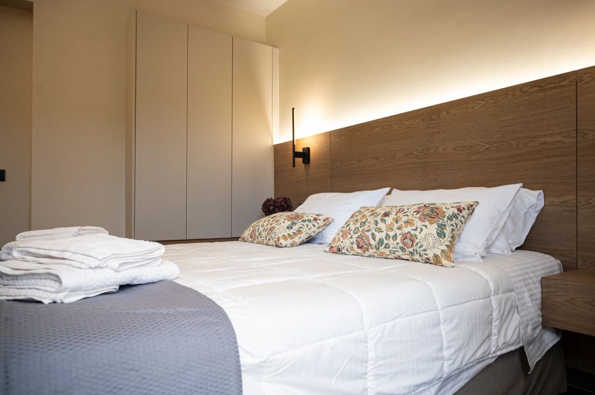 Bedroom with a double bed, patterned cushions, towels and a wooden headboard with indirect lighting