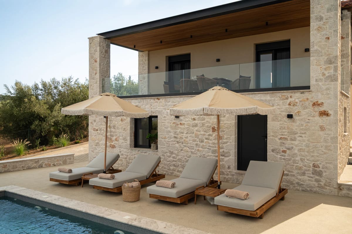 The two-storey stone house with a glass balcony, the terrace with loungers and parasols, and the pool in front