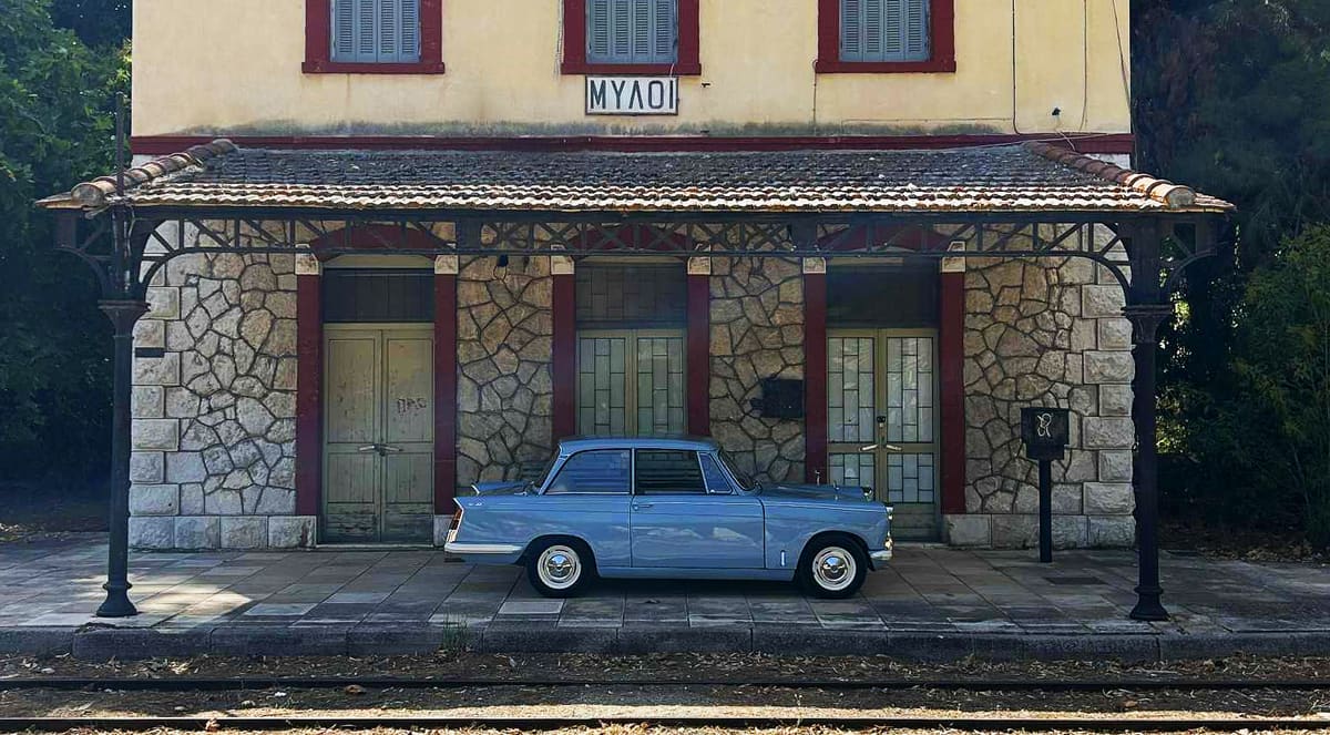 A blue 1960s Triumph Herald parked in front of the old Myloi railway station
